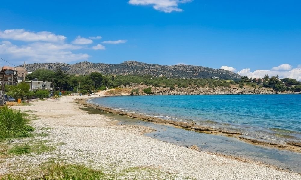 Παραλία Καλός Γιαλός στο Πόρτο Ράφτη με ήρεμα γαλάζια νερά, βοτσαλωτή ακτή, χαμηλή βλάστηση και θέα στους λόφους της ανατολικής Αττικής.